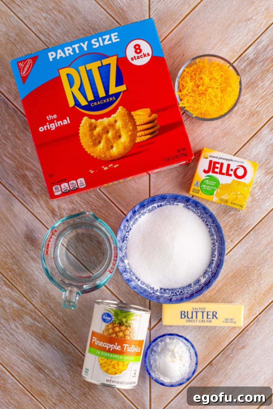 A vibrant flat lay of all the essential ingredients for pineapple casserole: Ritz crackers, pineapple gelatin mix, shredded sharp cheddar cheese, sugar, cornstarch, and canned pineapple tidbits.