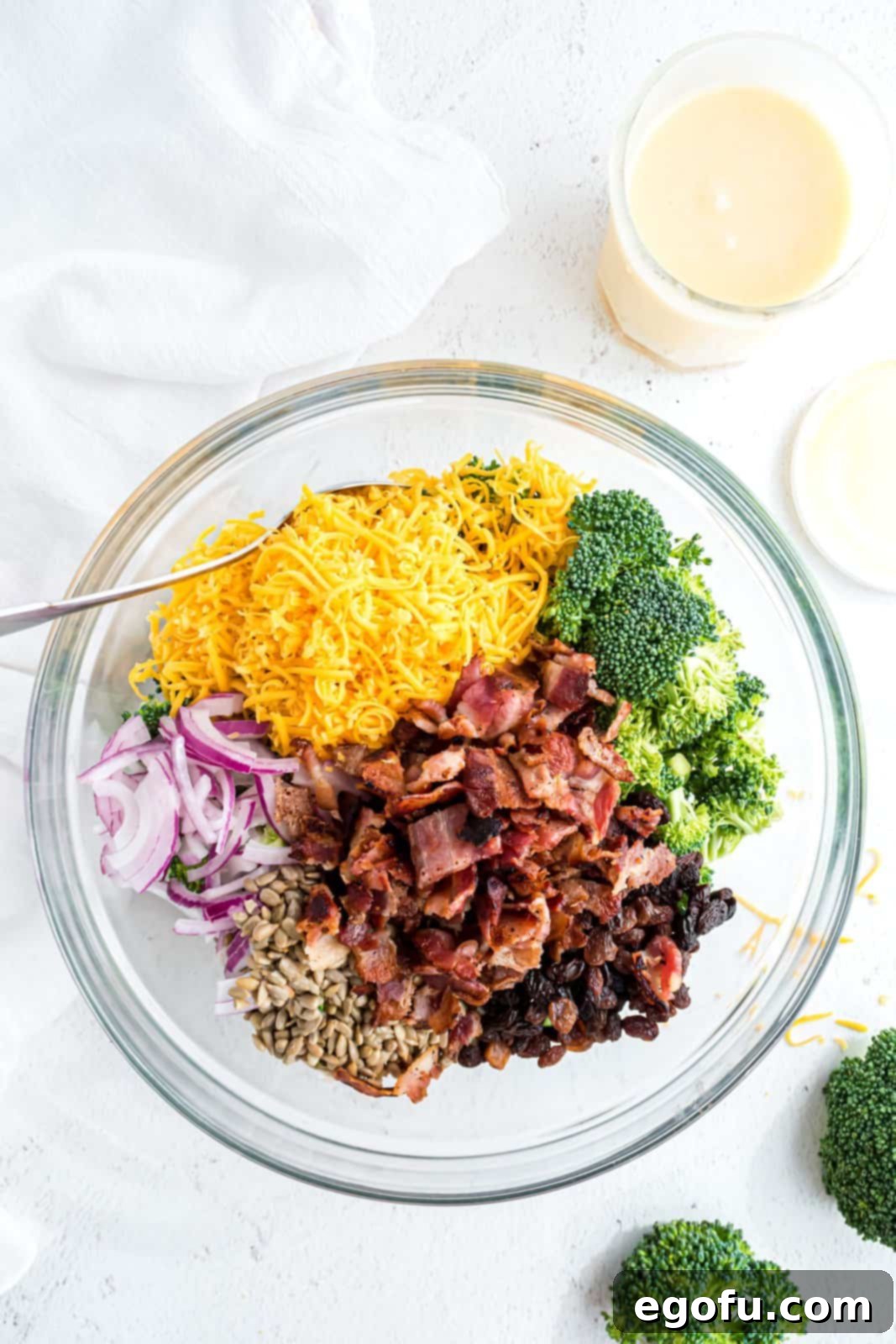 A large bowl containing all the dry salad ingredients: broccoli, bacon, raisins, walnuts, onions, sunflower seeds, and cheese.