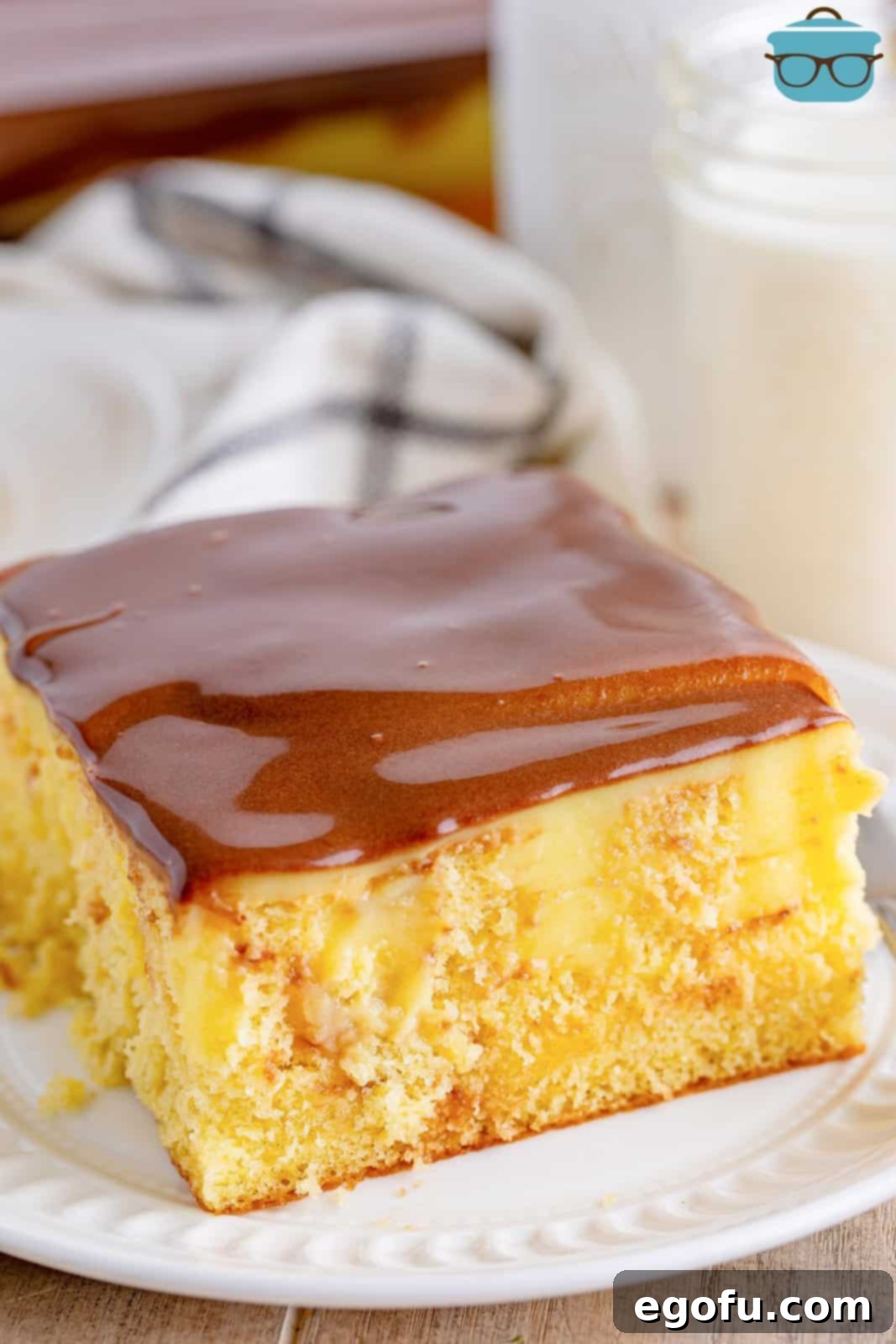 A delectable slice of Boston Cream Poke Cake, featuring distinct layers of moist yellow cake, creamy vanilla pudding, and a smooth, rich chocolate frosting, served elegantly on a small round white plate with a bottle of milk subtly blurred in the background, suggesting a perfect pairing.