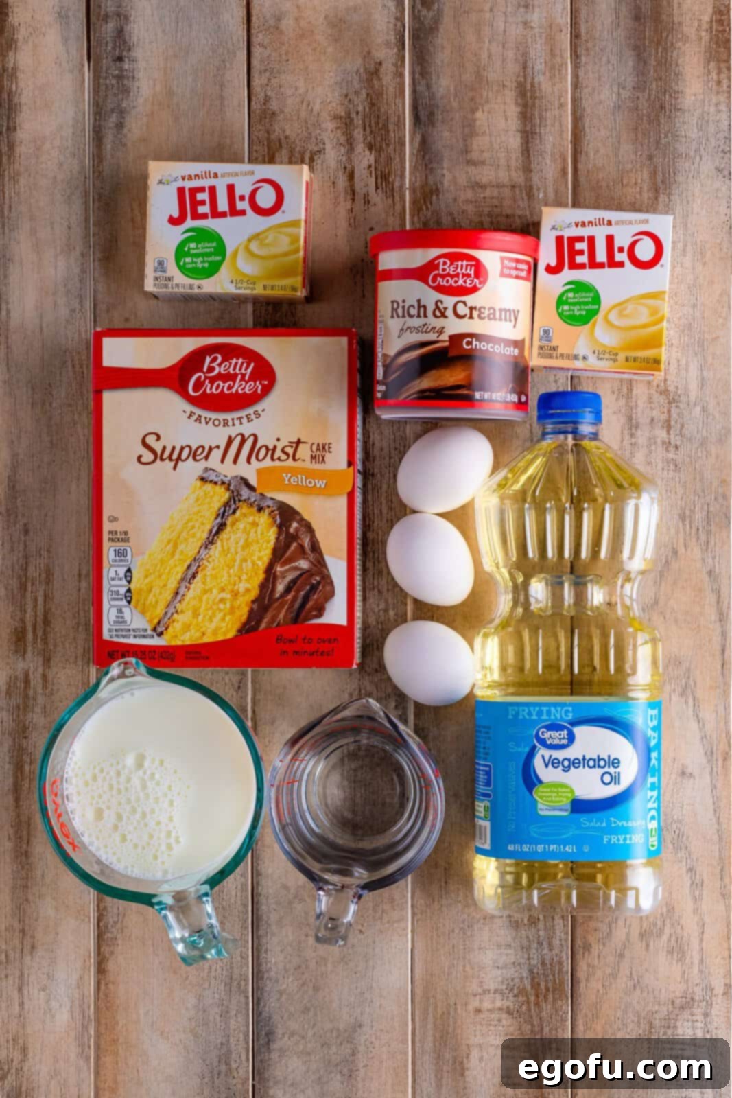 A beautifully arranged flat lay photograph displaying all the essential ingredients required for making Boston Cream Poke Cake: a box of yellow cake mix, two boxes of instant vanilla pudding mix, a tub of creamy chocolate frosting, fresh eggs, a bottle of cooking oil, a glass of water (or milk), and a carton of milk, neatly organized on a clean kitchen surface.