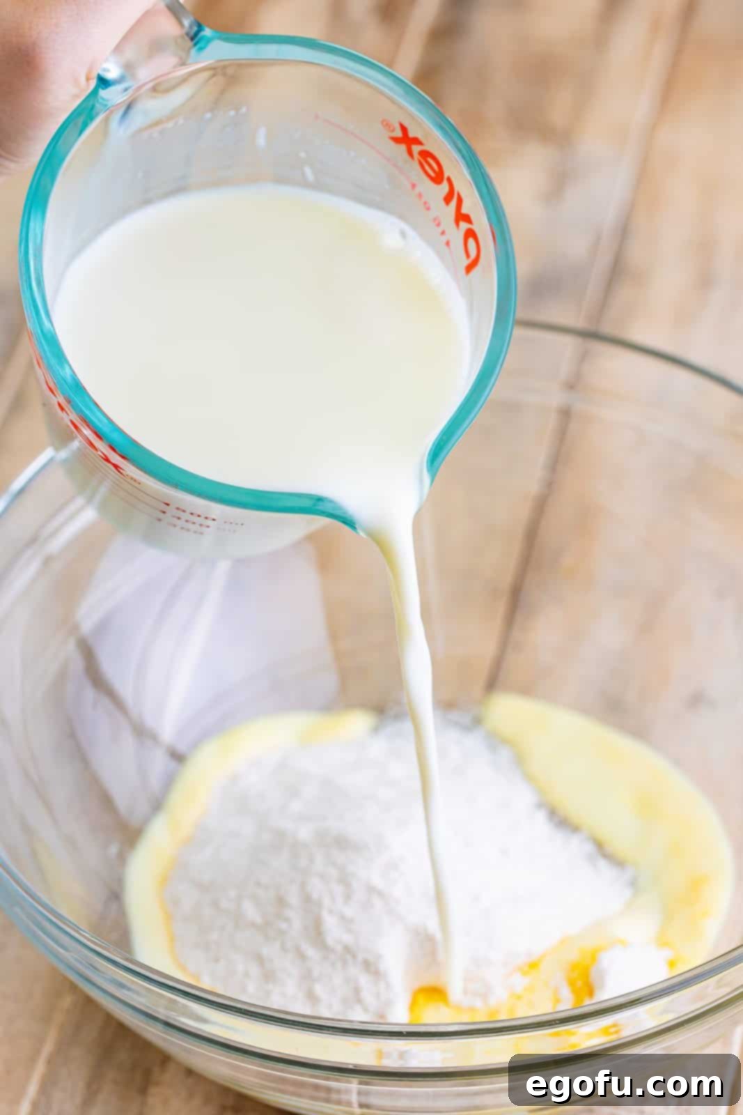 A clear glass mixing bowl prominently displaying instant vanilla pudding mix as a stream of cold milk is poured directly on top, illustrating the initial and crucial step of pudding preparation for the poke cake.