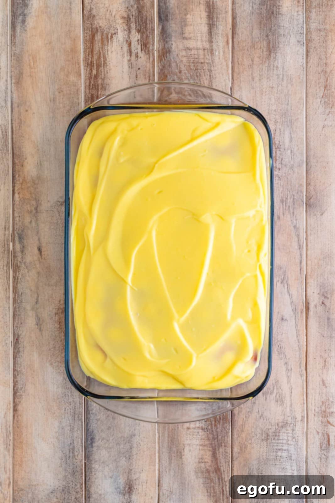 A delicious and inviting layer of creamy vanilla pudding spread smoothly and evenly across the entire top surface of a baked yellow cake, effectively filling all the previously poked holes and creating a seamless, uniformly creamy layer, ready for chilling.