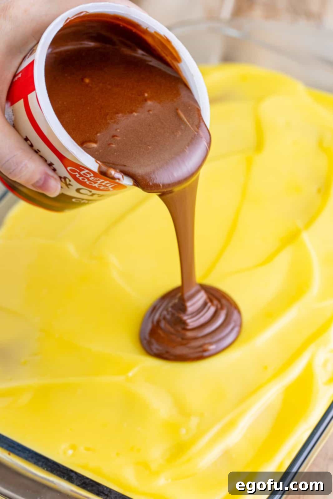 A delightful moment captured in progress, as smooth, melted chocolate frosting is being poured generously over a creamy vanilla pudding layer, which in turn rests atop a moist yellow cake base, creating the final decadent and iconic layer of the Boston Cream Poke Cake.