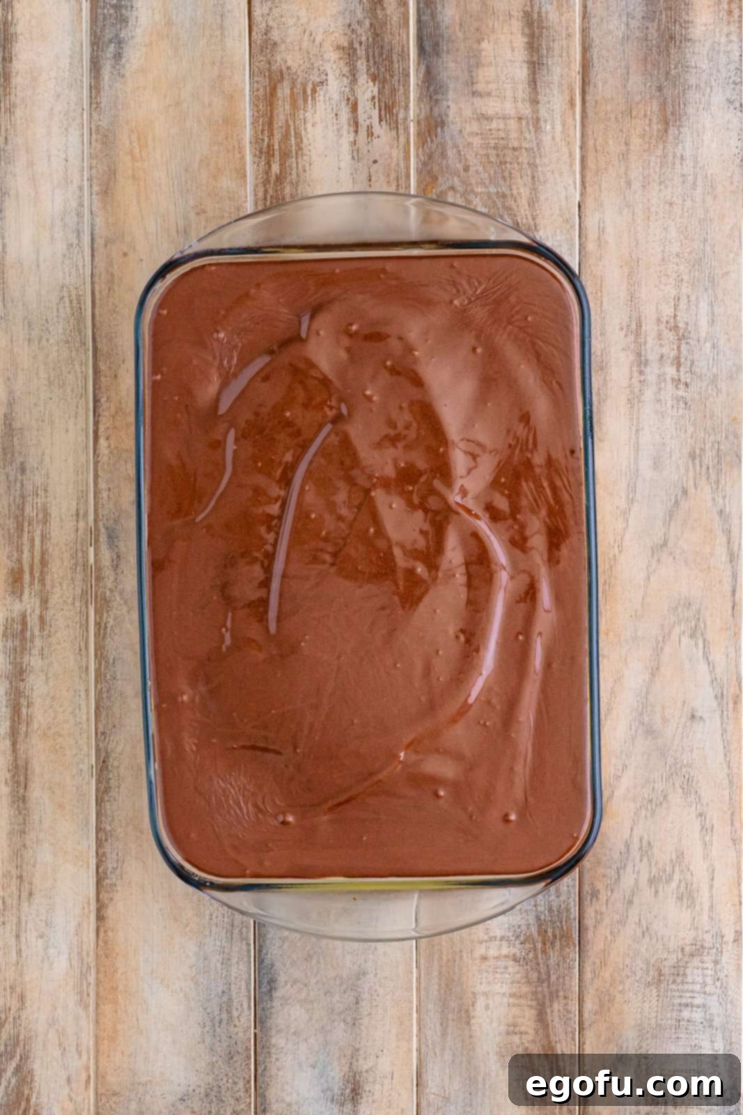 A pristine, fully frosted Boston Cream Poke Cake, presented as an overhead view, showcasing its perfectly smooth, even, and glistening chocolate topping, with the edges neatly covered, indicating it's ready to be chilled and enjoyed.