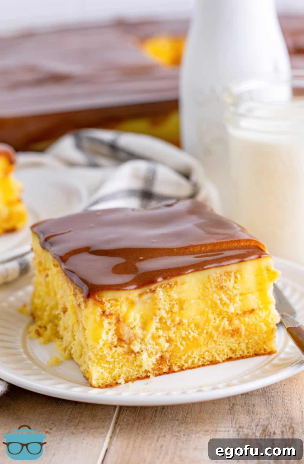 A beautifully presented Boston Cream Poke Cake showcased on a pristine white round plate, with a vintage-style glass bottle of milk and the original baking pan subtly blurred in the background, creating a rustic, inviting, and tempting dessert scene.