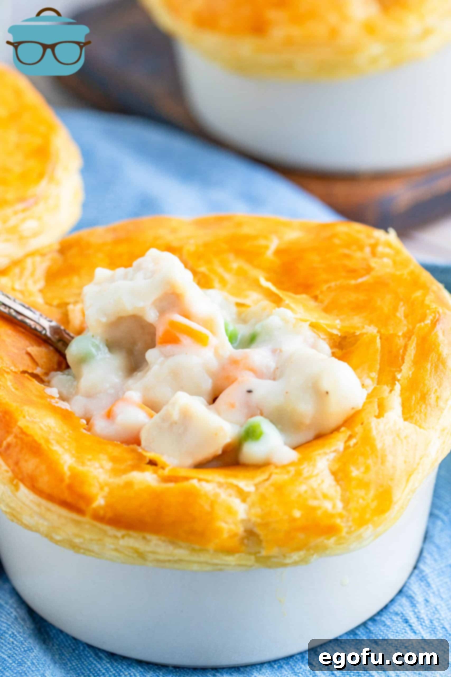 Closeup of a golden-brown individual chicken pot pie in a white ramekin, with a spoon gently breaking into the flaky puff pastry crust and creamy filling visible below.