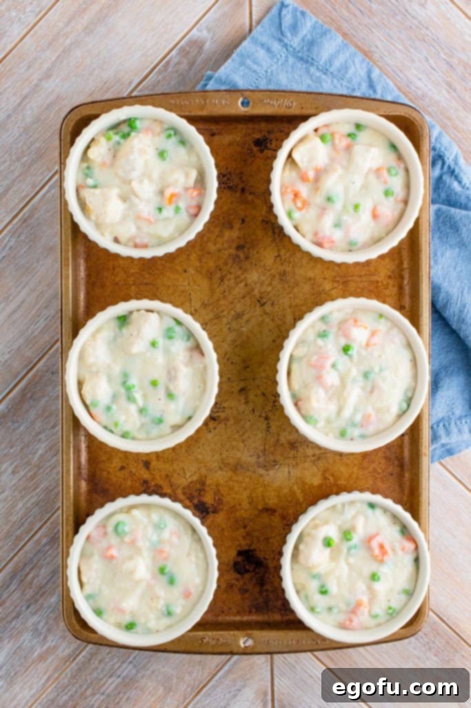 Six ramekins filled about three-quarters full with creamy chicken pot pie filling, placed on a baking sheet, ready for the crust.