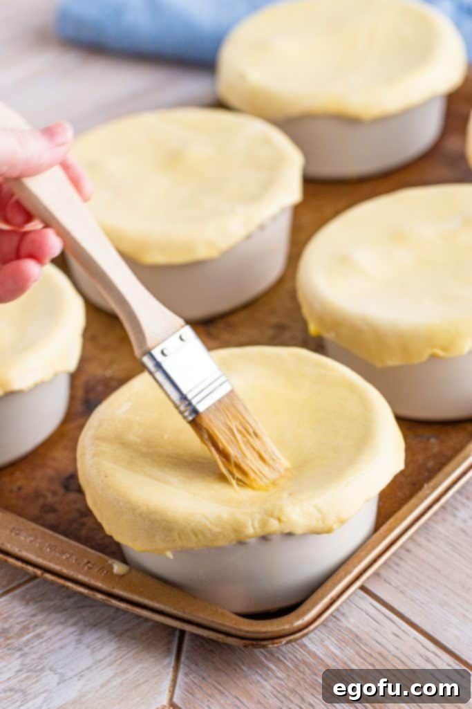 A pastry brush applying egg wash to the puff pastry tops of individual chicken pot pies in ramekins before baking.