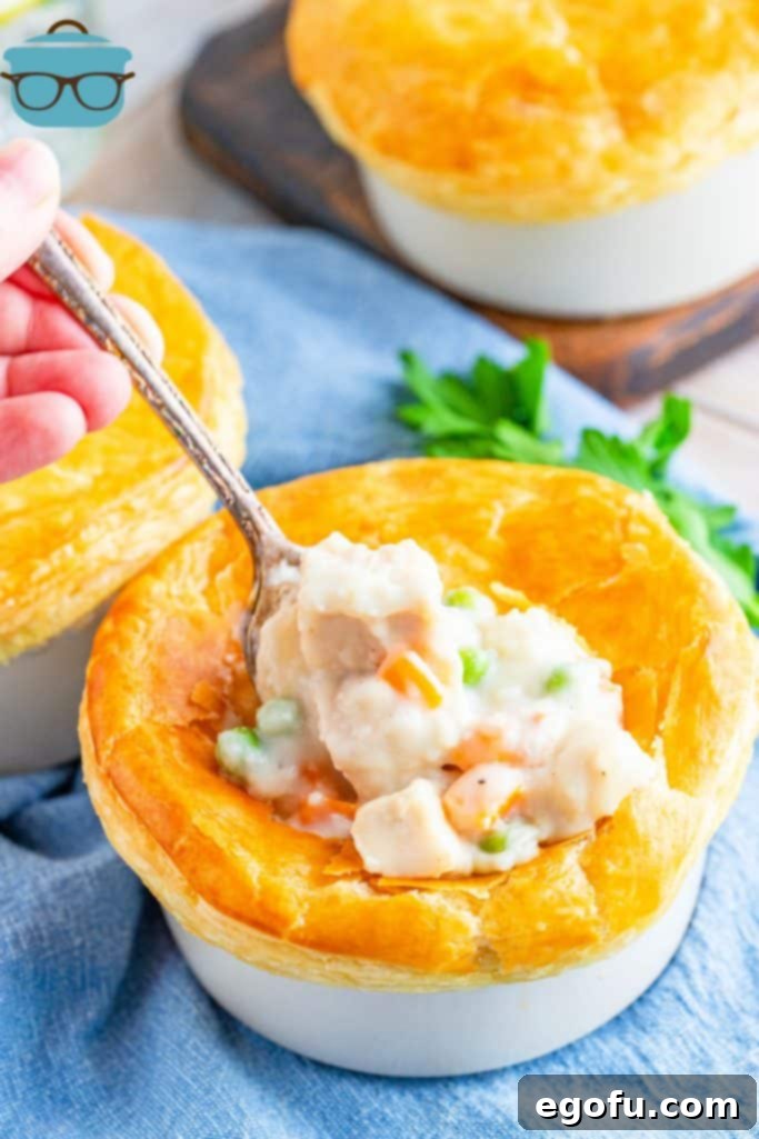 A spoon inserted into a baked individual chicken pot pie, revealing the creamy chicken and vegetable filling beneath the golden puff pastry crust.