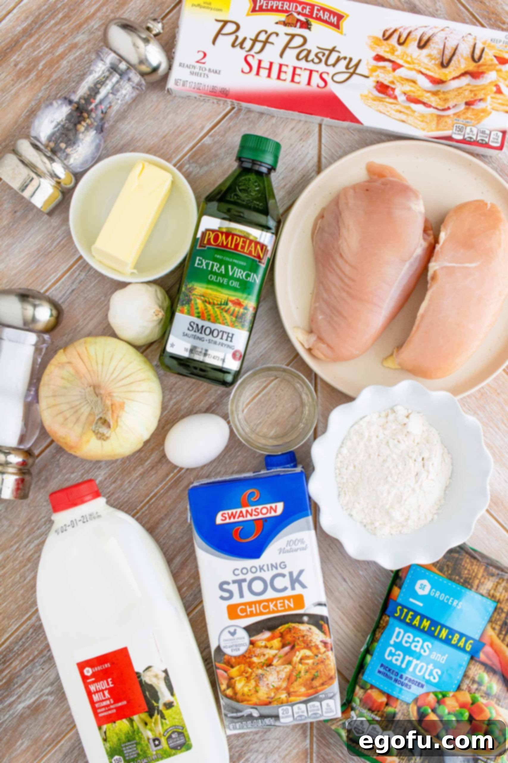 A flat lay image displaying all the ingredients for Homemade Chicken Pot Pie: frozen puff pastry, raw chicken breasts, olive oil, salt and pepper, butter, flour, whole milk, chicken stock, diced onion, minced garlic, and a bag of frozen peas and carrots.