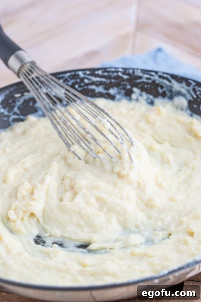 Whole milk being slowly added to the roux in a pan, with a whisk stirring to combine the mixture.
