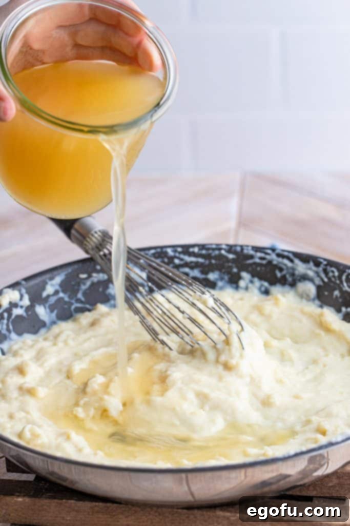 Chicken stock being poured into the creamy milk and roux mixture in a large pan, alongside chopped onion and garlic, while being whisked.