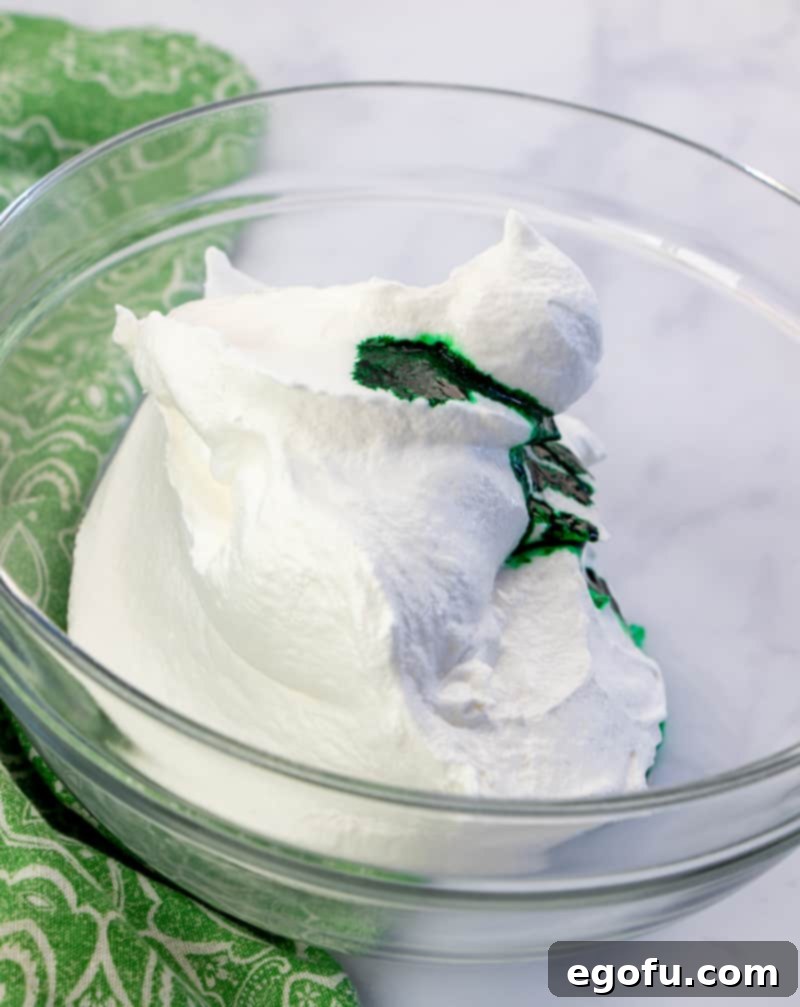 Effortless Grasshopper Indulgence 9 Thawed whipped topping (Cool Whip) being mixed with mint extract and green food coloring in a bowl, creating a light green, minty topping.
