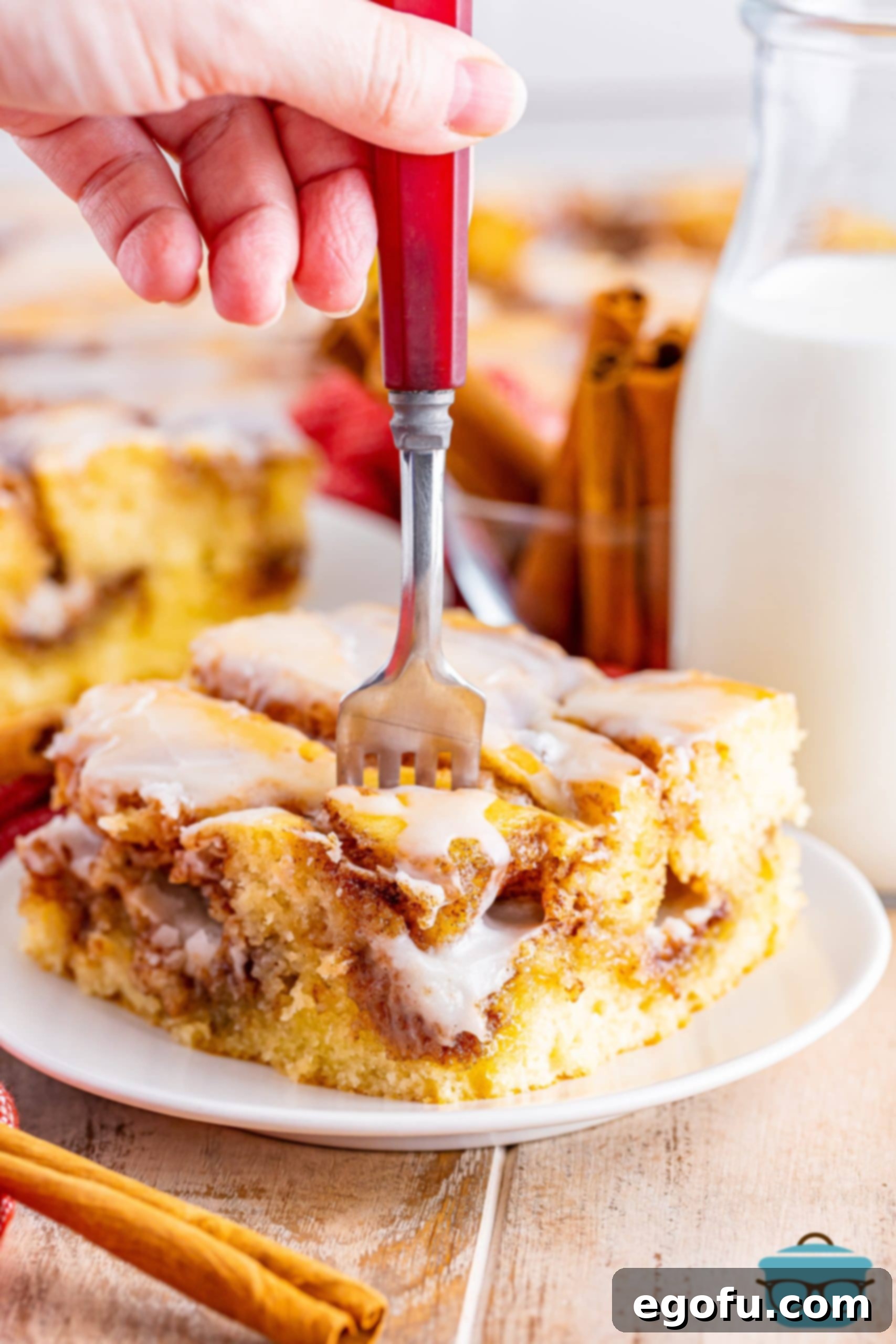 Swirl of Spice Cake 15 A fork is gently inserted into a rich, glazed slice of Cinnamon Roll Cake on a pristine white plate, inviting a taste.