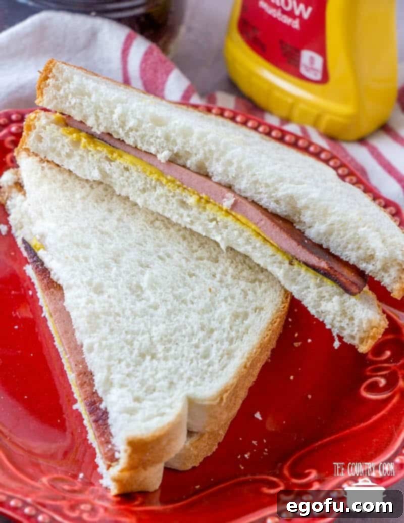 Fried bologna on soft white bread with mustard, sliced in half on a red plate, ready to be enjoyed as a classic comfort food sandwich.
