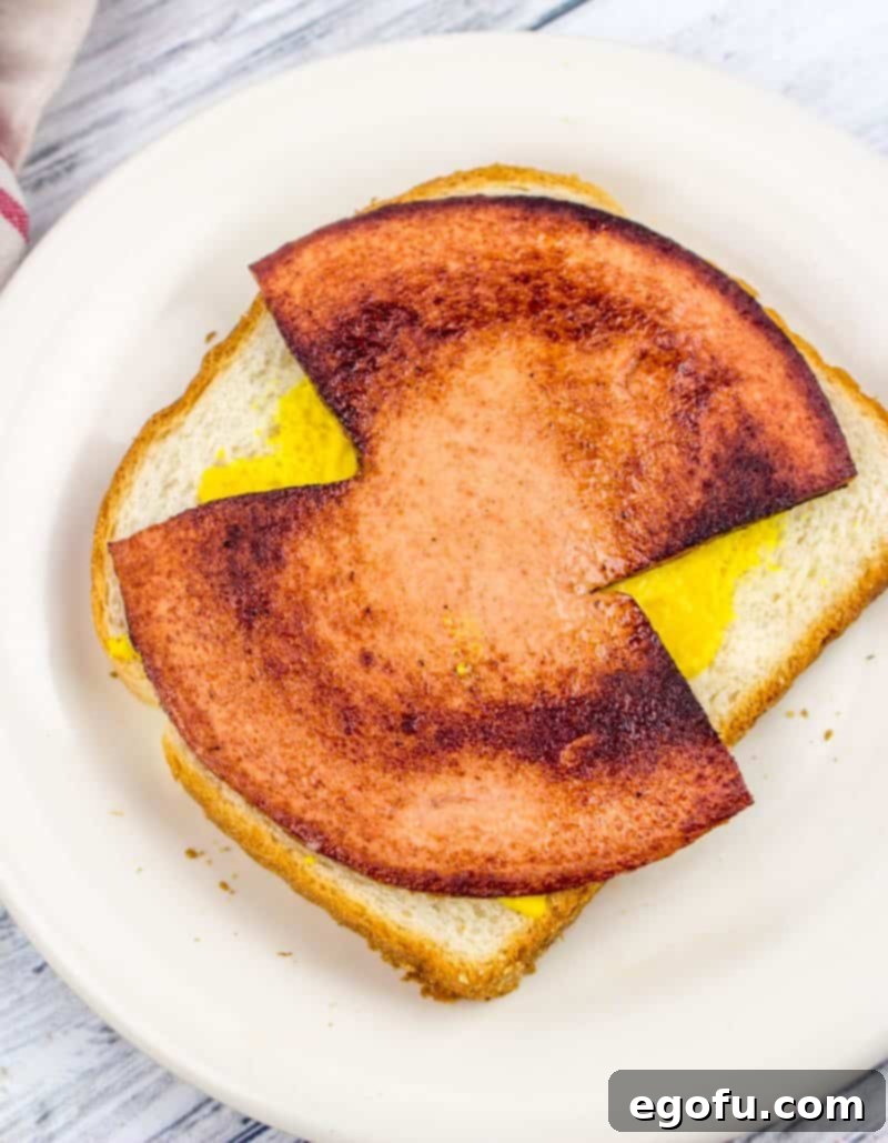 Crisp fried bologna resting on a slice of soft white bread coated with yellow mustard, forming the core of a simple, satisfying sandwich.