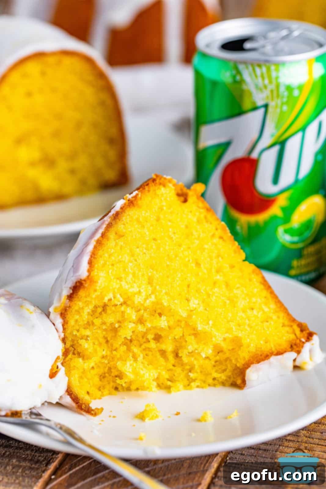A perfectly sliced piece of 7Up cake resting on a small, round white plate. A fork has just taken a small bite from the cake, revealing its moist, tender interior. The plate sits on a rustic wooden surface, highlighting the cake's inviting texture.