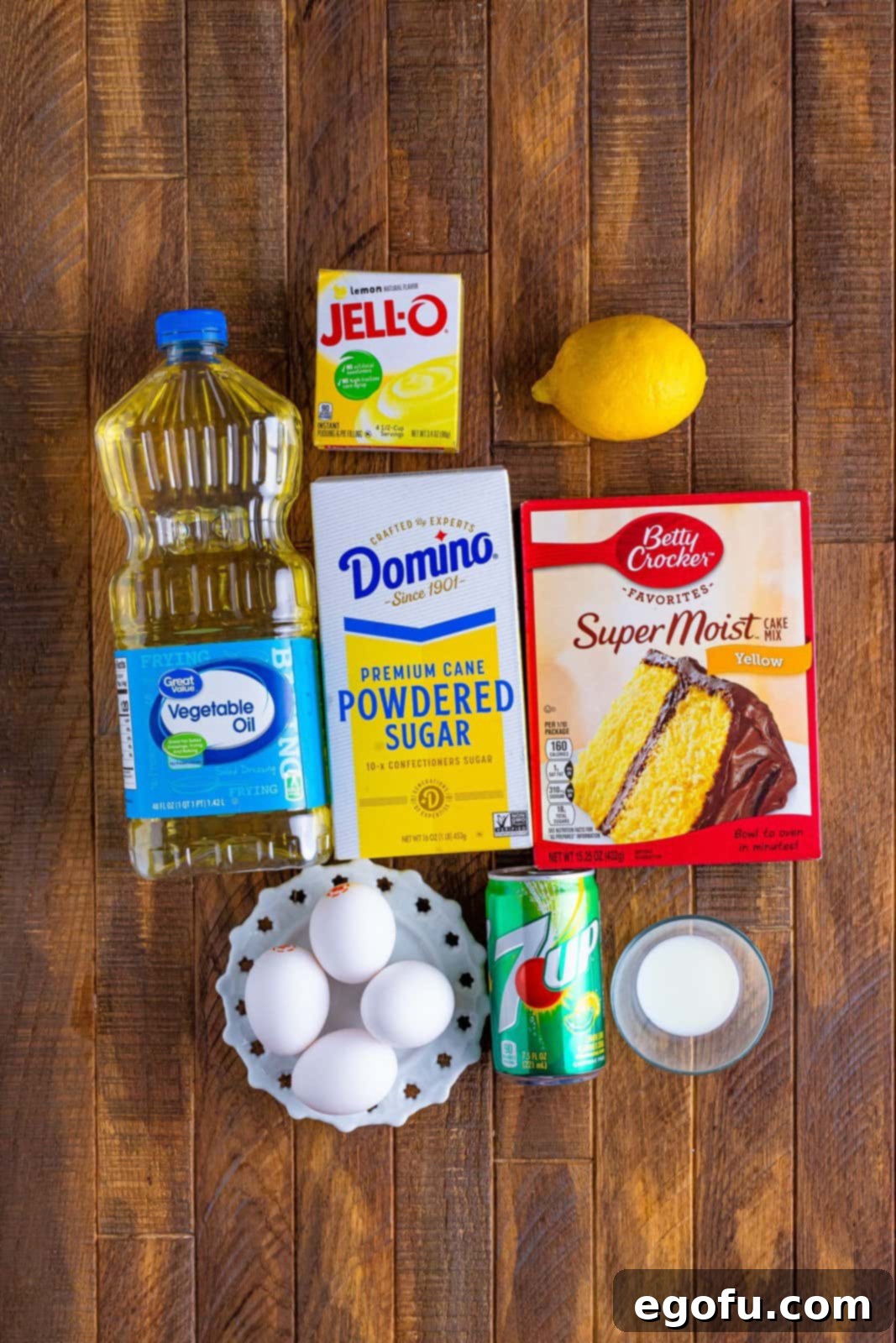 A vibrant flat lay shot displaying all the essential ingredients for a 7Up Bundt Cake: a box of yellow cake mix, a box of instant lemon pudding, a can of 7Up soda, fresh large eggs, a bottle of vegetable oil, and a bright yellow lemon, with a container of sour cream also present, though not used in the final version of this recipe.