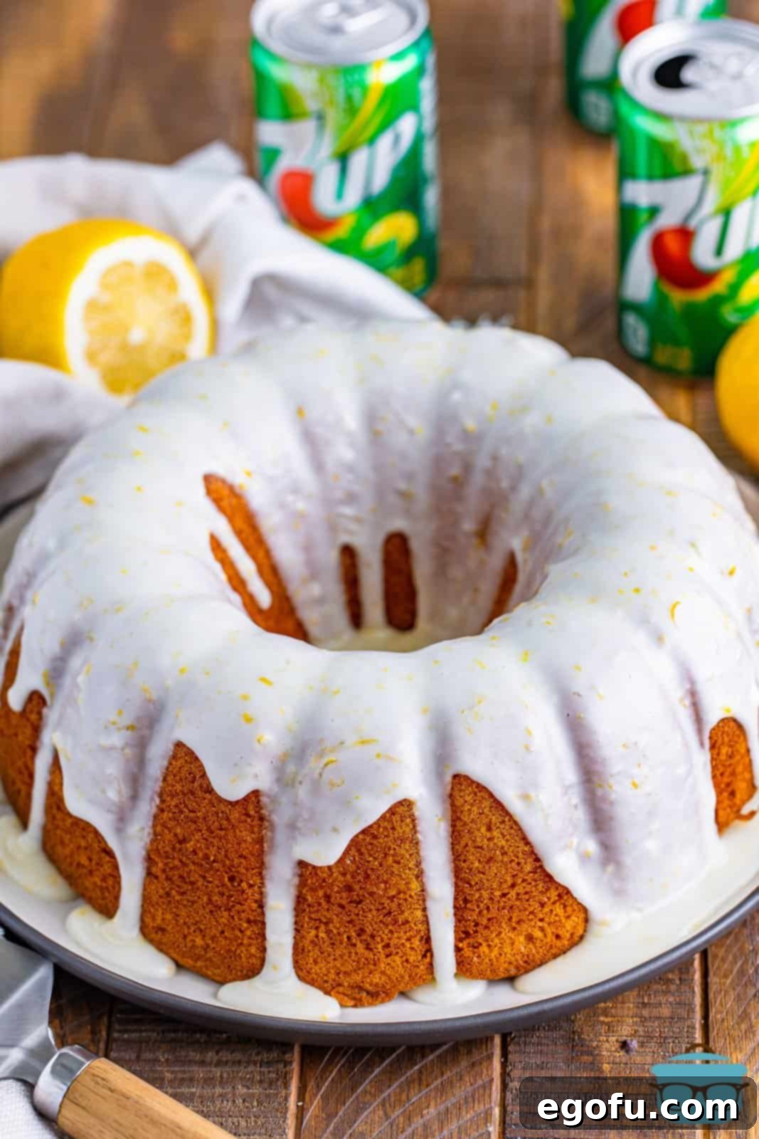 A breathtaking final shot of the fully baked 7Up cake, elegantly presented on a white cake plate. A generous drizzle of bright lemon icing cascades over its golden surface, while several cans of 7Up stand prominently in the background, reinforcing the cake's signature ingredient. The image perfectly captures the cake's fresh, inviting appeal.