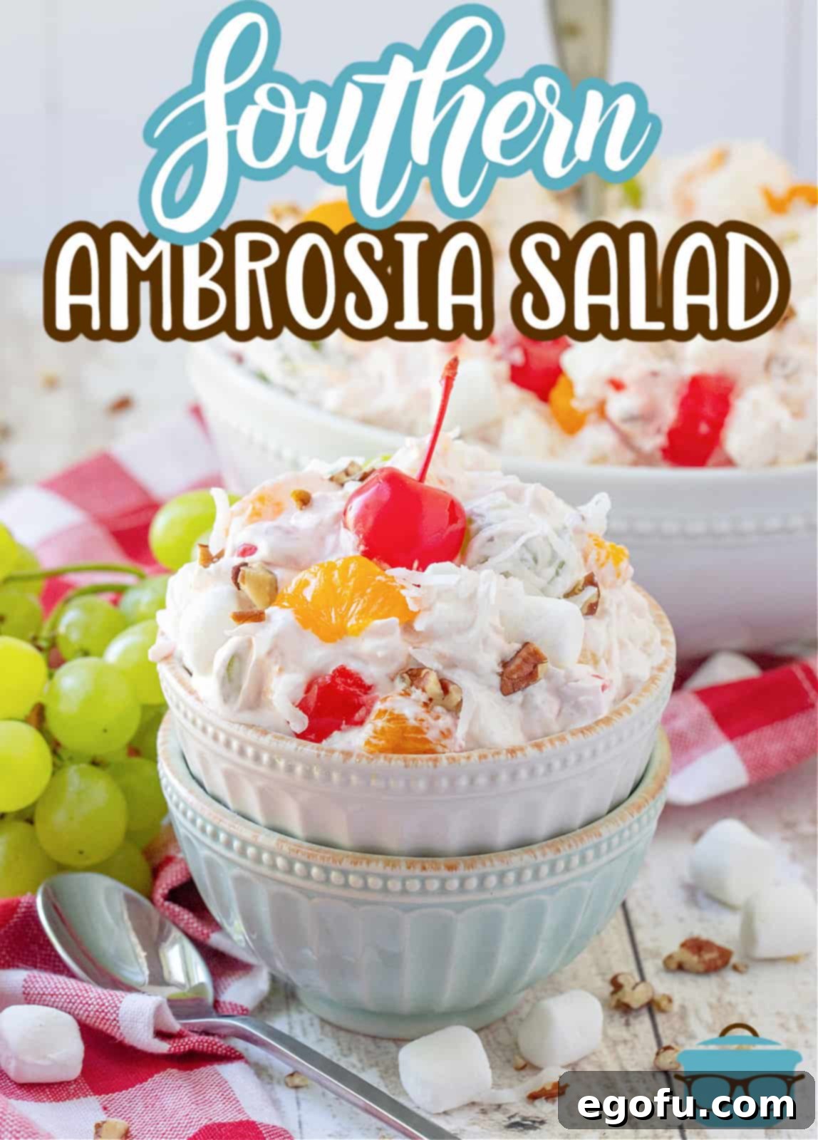 Southern Ambrosia Delight 3 Southern Ambrosia Salad recipe from The Country Cook, a white and blue bowl stacked on top of each other, a serving of ambrosia salad shown in top bowl with a maraschino cherry on top and a bunch of green grapes in the background.