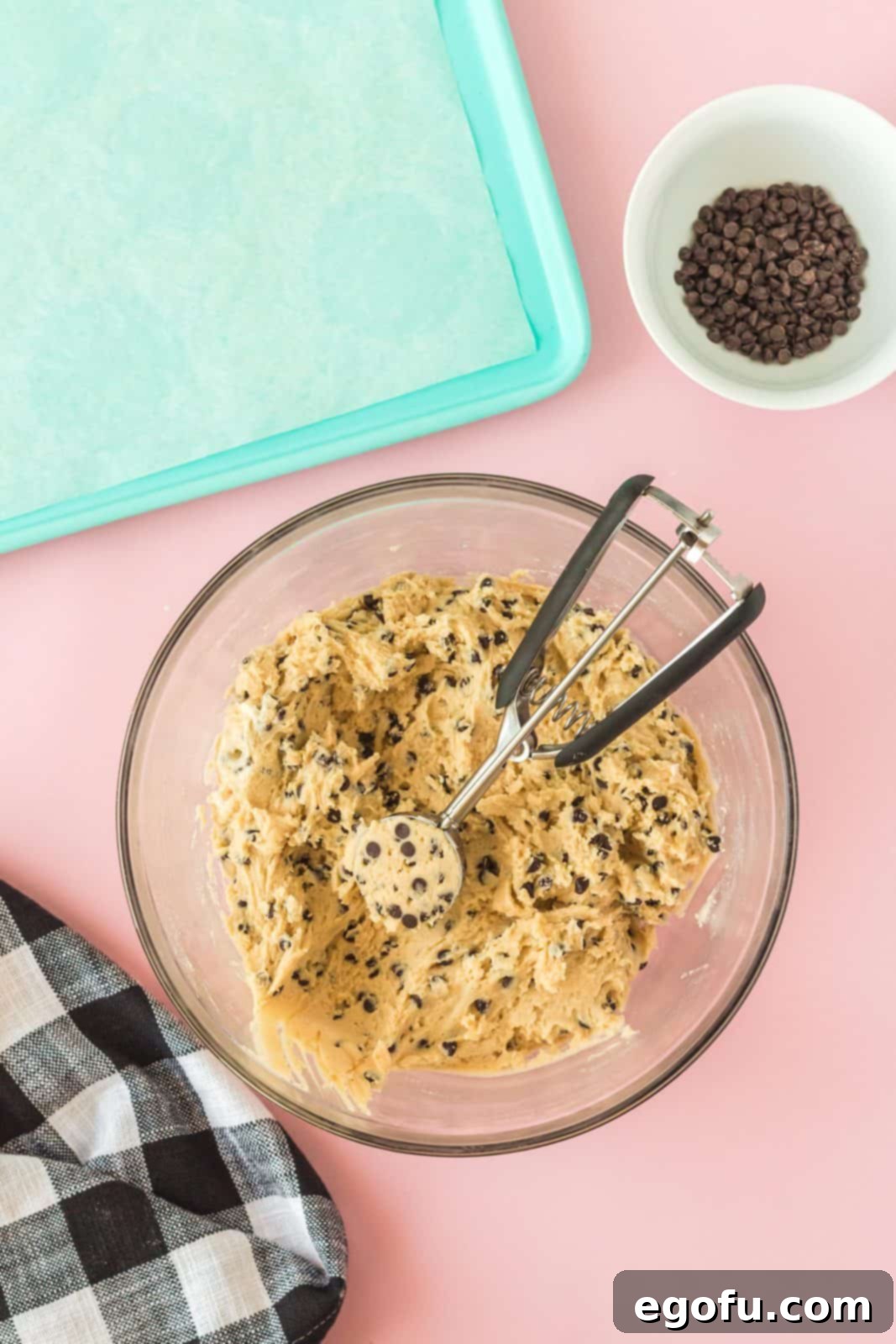 The flour mixture being added to the butter and cream cheese mixture, with cookie dough already scooped onto a baking sheet in the background, showing chocolate chips folded in.