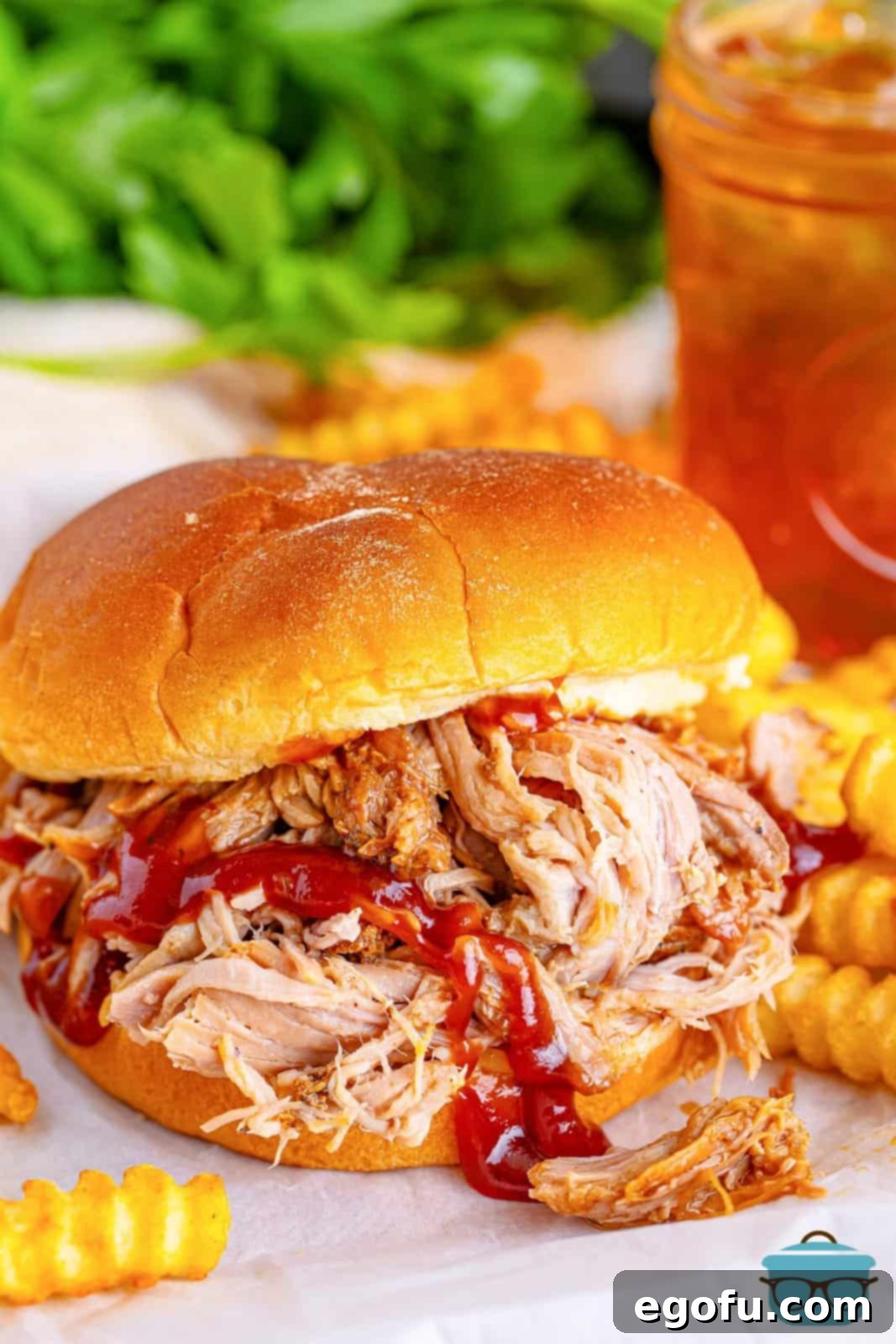 A pulled pork BBQ sandwich on white parchment paper, surrounded by scattered crinkle-cut French fries.