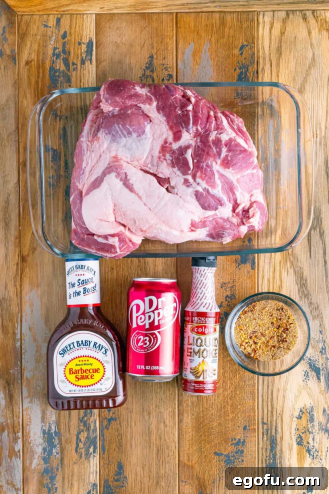 Pork shoulder roast, BBQ sauce, a can of Dr Pepper, hickory liquid smoke, and a bowl of pork seasoning laid out on a surface.