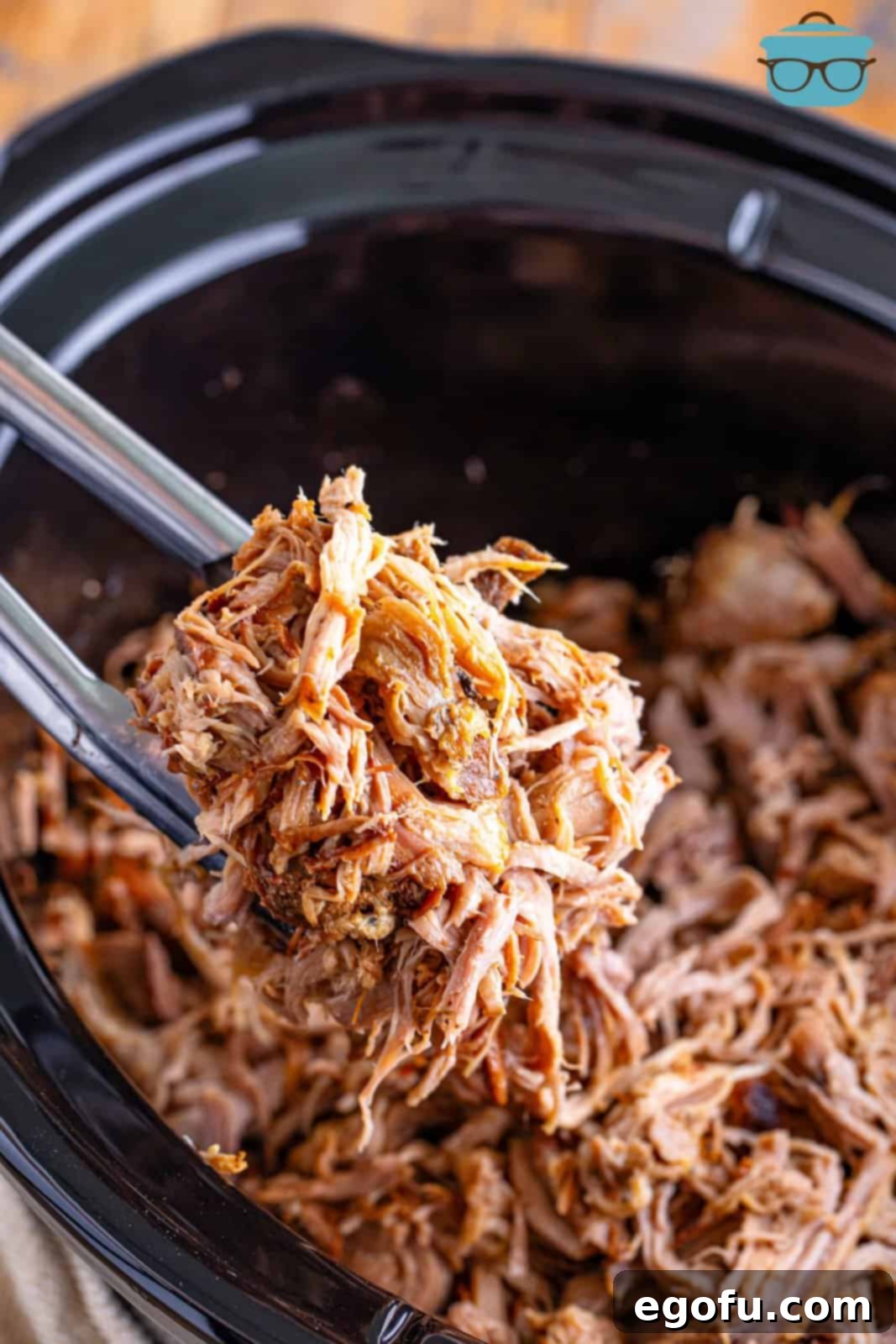 A pair of tongs holding up a generous portion of shredded pulled pork over the slow cooker.