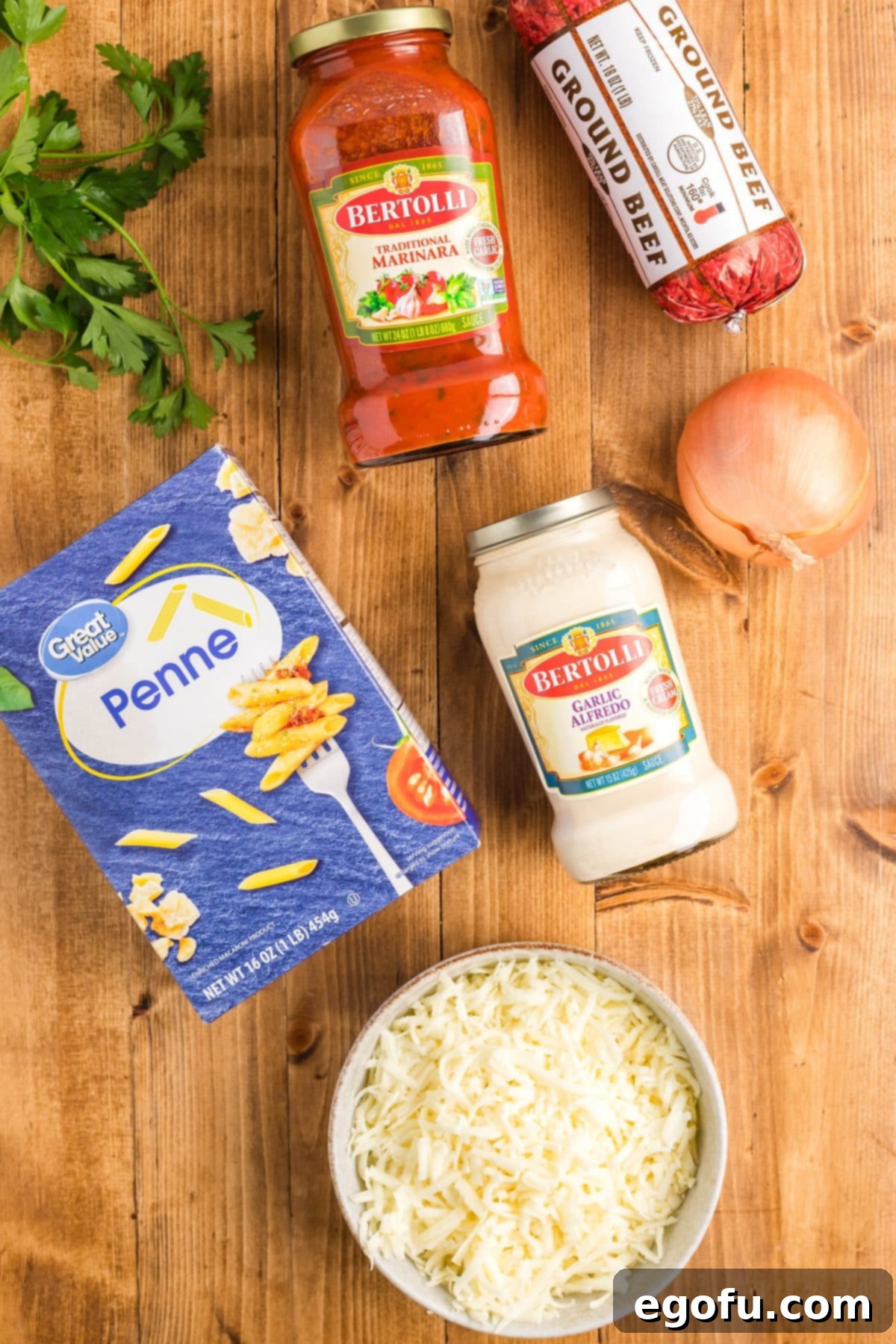 A flat lay photograph showcasing the primary ingredients for baked ziti: uncooked penne pasta, a jar of garlic alfredo sauce, a whole yellow onion, raw ground beef, a jar of spaghetti sauce, and a bowl of shredded mozzarella cheese.