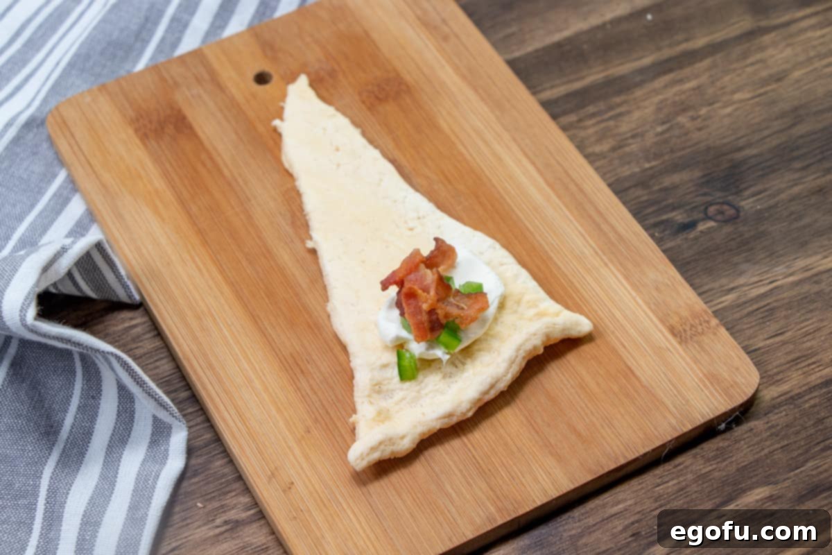 Zesty Jalapeño Delights 7 A crescent roll triangle, topped with a generous dollop of cream cheese, a sprinkling of diced jalapeños, and crispy bacon bits, ready to be rolled.