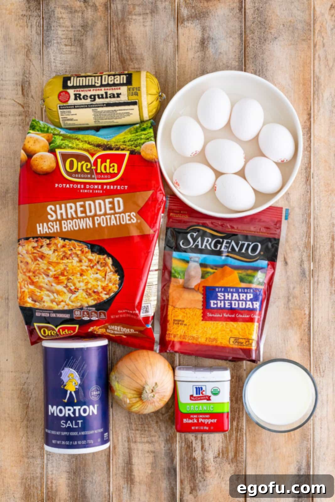 A flat lay of the key ingredients: pork sausage, hash browns, eggs, shredded cheddar cheese, salt, pepper, and onion, laid out on a wooden surface.