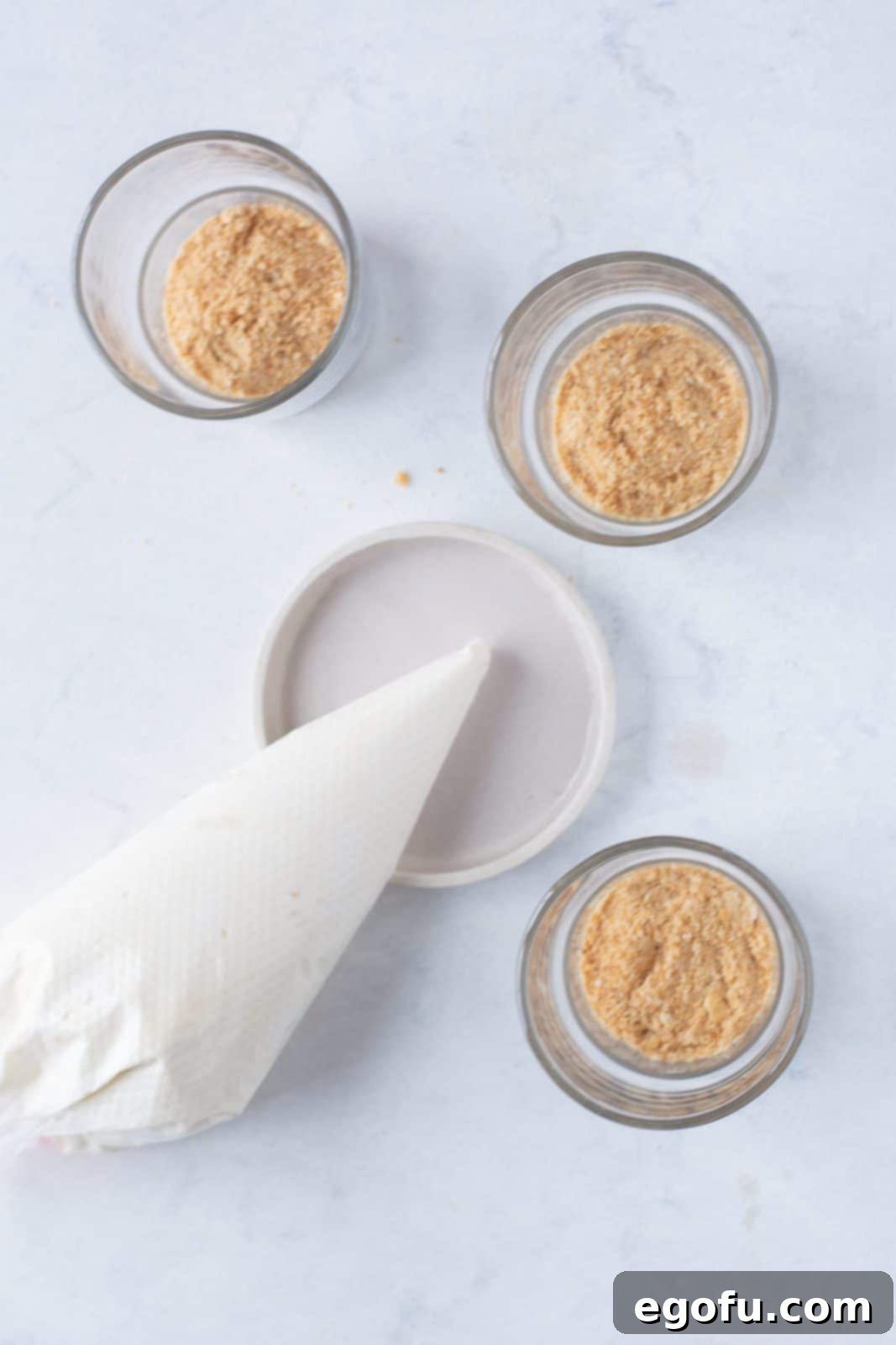 No-Bake Strawberry Cheesecake Parfaits 8 A piping bag filled with the creamy cheesecake mixture, strategically placed next to the prepared glasses that already contain the graham cracker crumb base, awaiting the next layer.