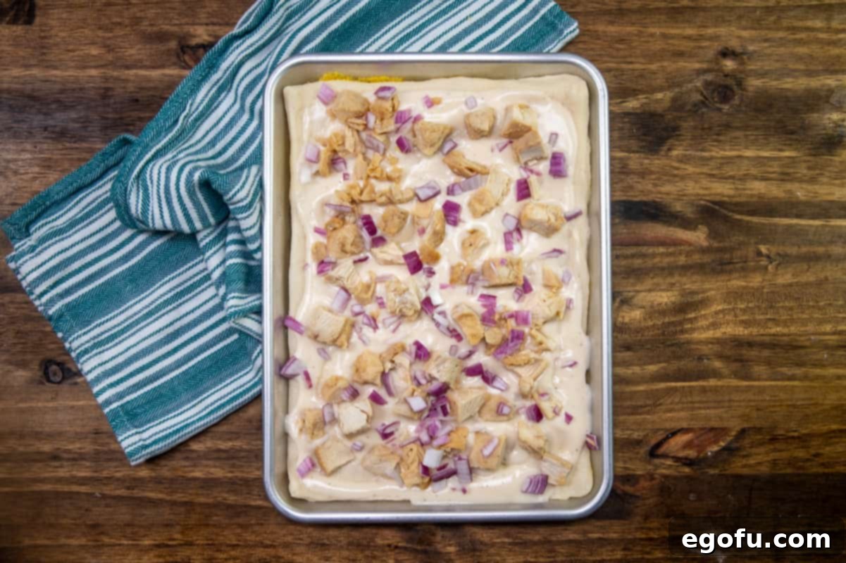 Diced grilled chicken and diced red onion spread out evenly on top of Alfredo sauce on pizza dough.
