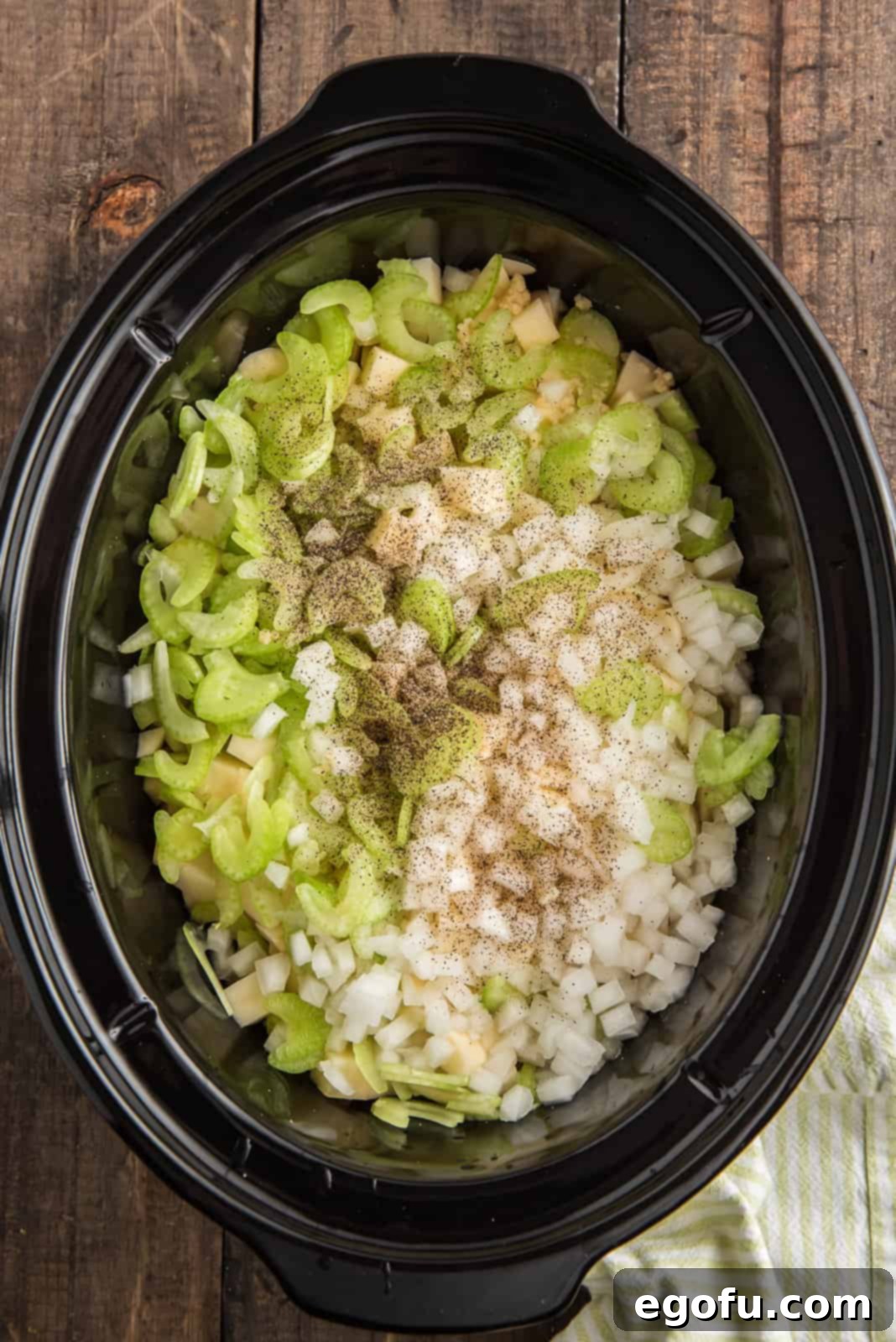 Slow Cooker Creamy Potato Bliss 4 Diced celery, chopped onion, minced garlic, salt, pepper, and rich chicken broth are added over a bed of diced potatoes in a slow cooker, illustrating the layering process before cooking.