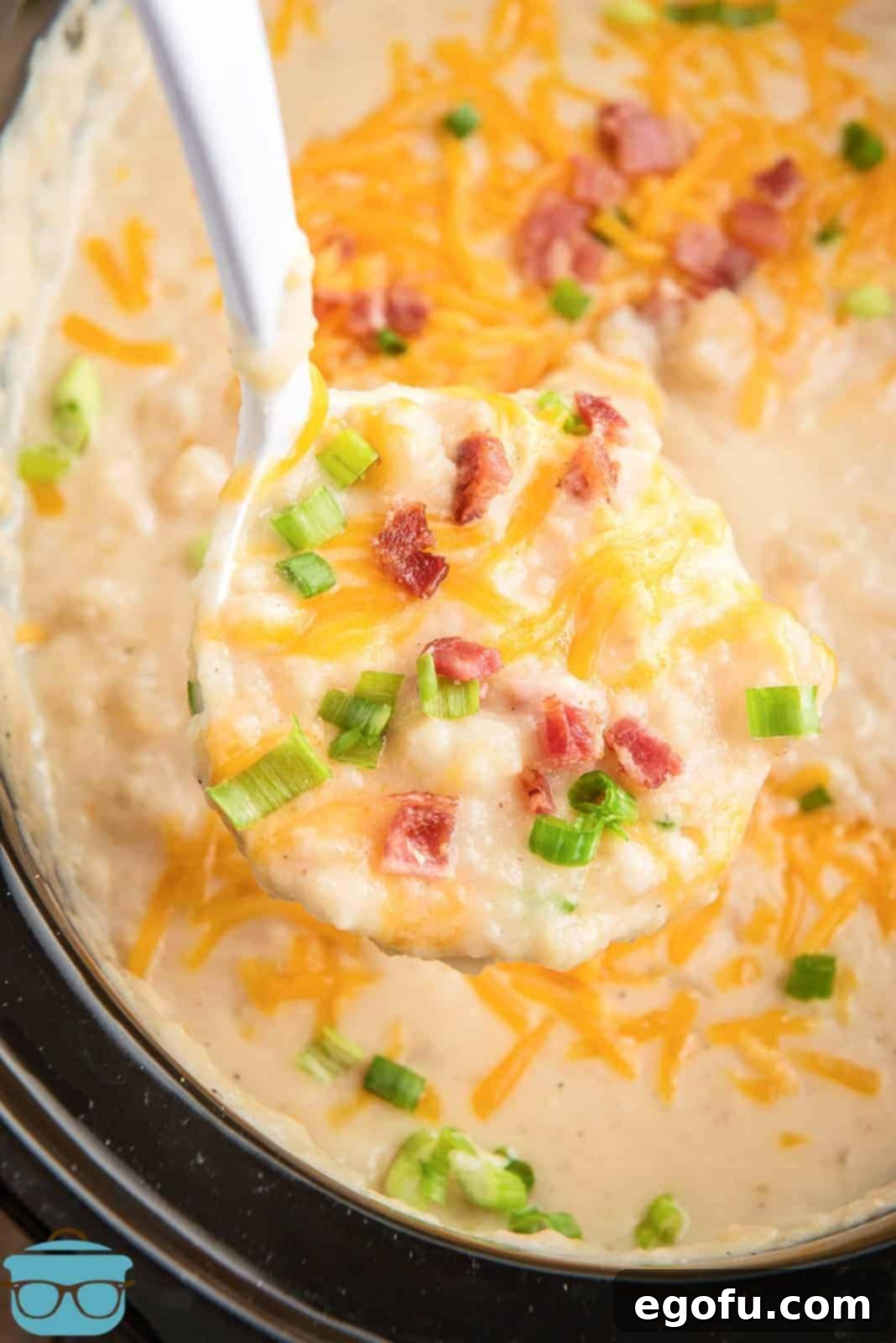 Slow Cooker Creamy Potato Bliss 9 A white bowl of creamy potato soup adorned with a generous topping of shredded cheddar cheese, crispy bacon bits, and fresh green onions.