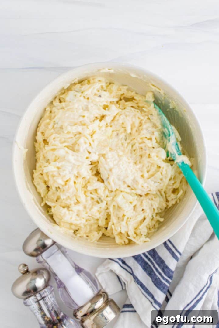 Thawed shredded hash browns and chopped onion being stirred into the sour cream mixture in a white bowl with a blue spoon, ensuring even coating.
