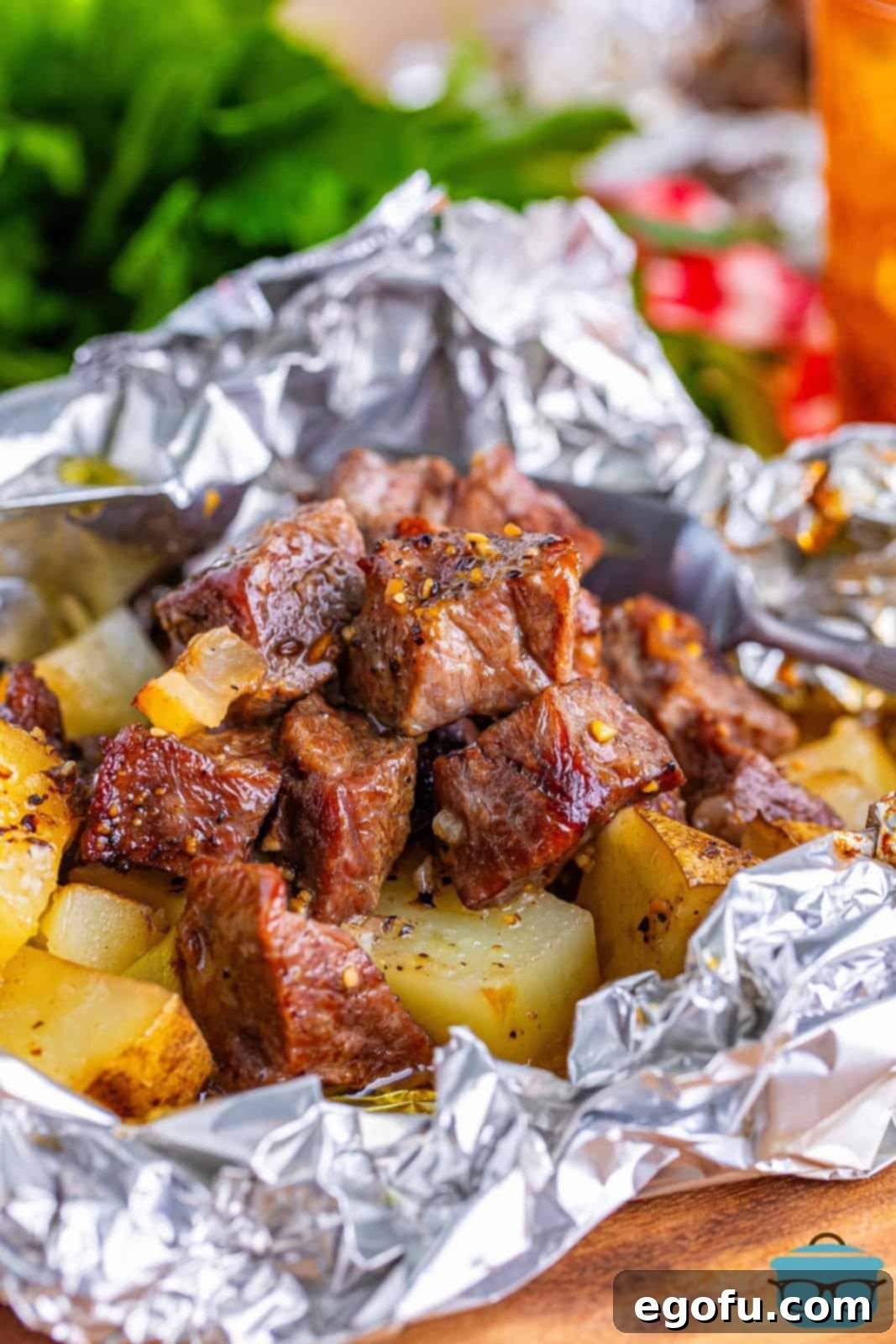an overhead view of grilled steak and potatoes in a crinkled foil packet with a glass of iced tea in the background. 