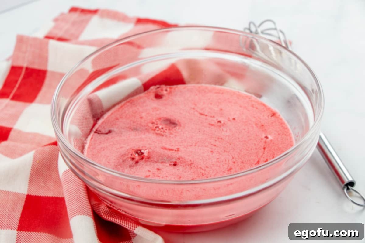 Strawberry Burst Poke Cupcakes 6 Strawberry gelatin powder being whisked into boiling water in a clear bowl, creating a smooth red liquid.
