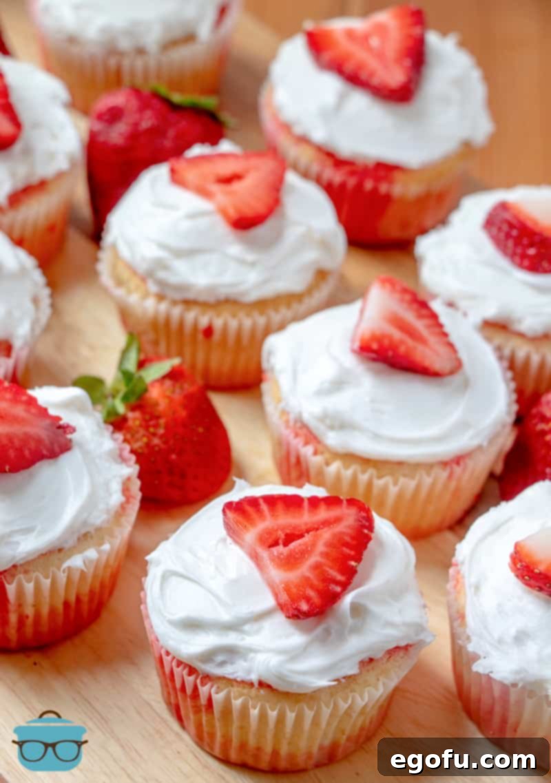 Strawberry Burst Poke Cupcakes 8 Several beautifully frosted Strawberry Jell-O Poke Cupcakes, topped with fresh strawberry slices, arranged on a rustic wooden cutting board.