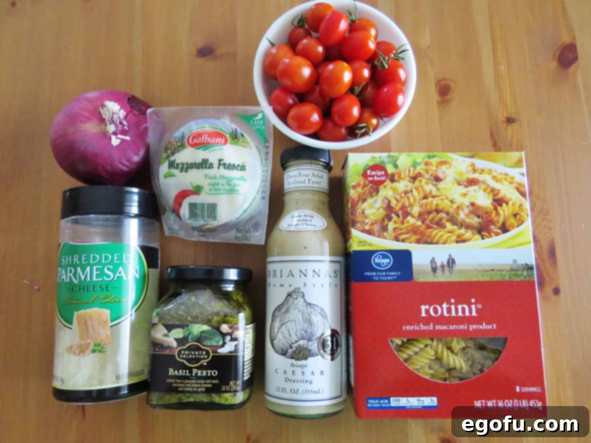 Individual ingredients laid out: Caesar salad dressing, basil pesto, fresh mozzarella, red onion, rotini pasta, and shredded Parmesan cheese.