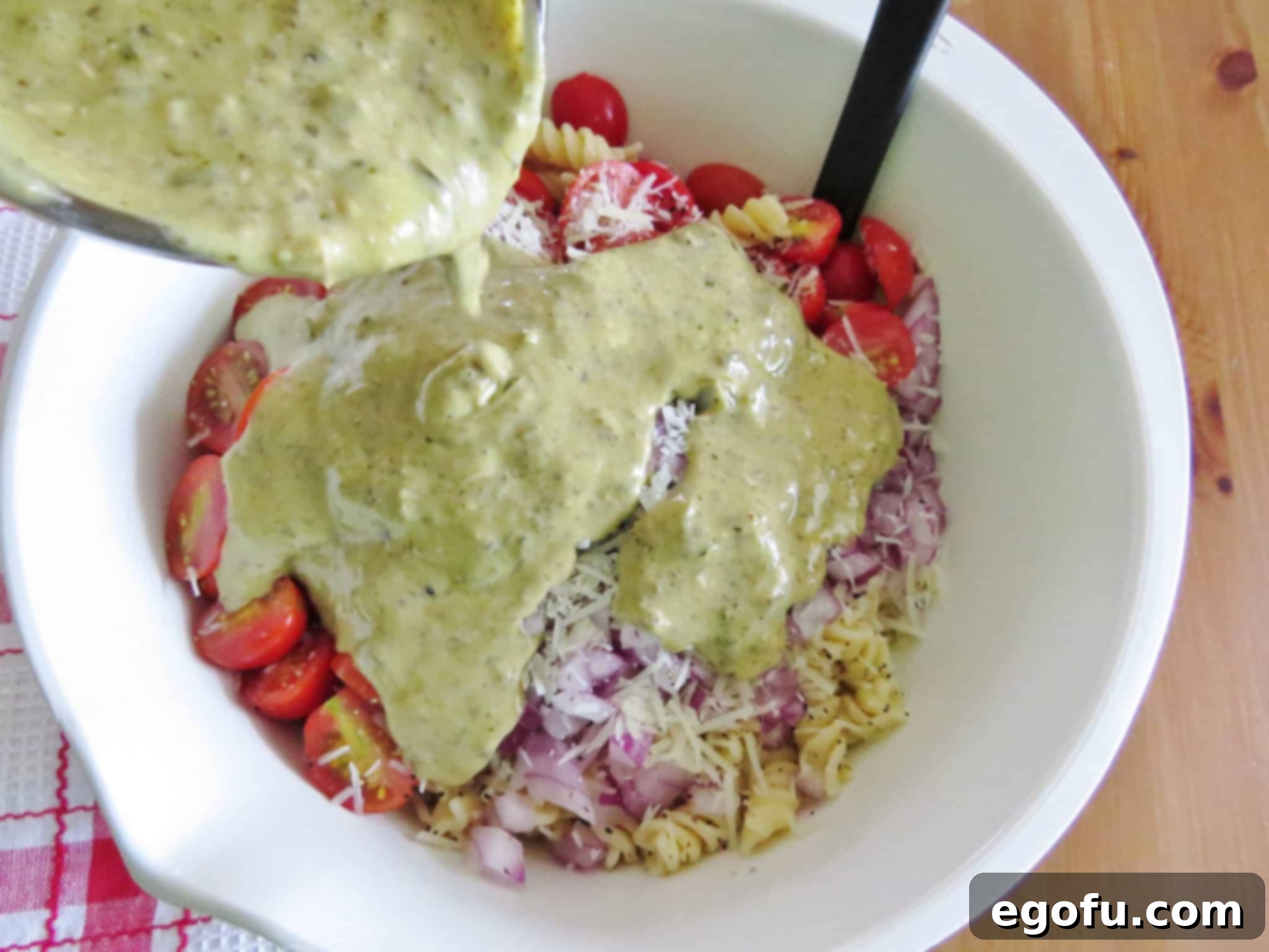 Sliced tomatoes, red onion, rotini pasta, and shredded Parmesan cheese in a large bowl, with Caesar basil pesto dressing freshly poured over them.