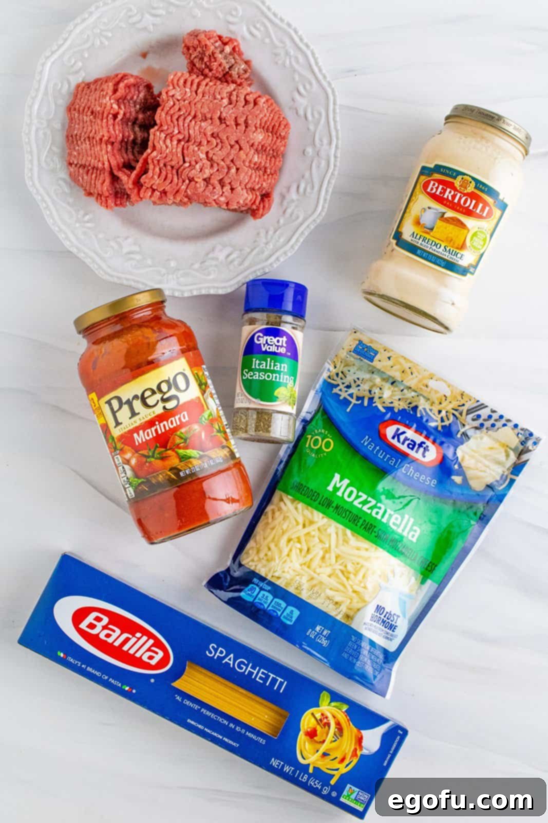Baked spaghetti ingredients laid out: spaghetti noodles, ground beef, spaghetti sauce, alfredo sauce (cheese sauce), Italian seasoning, and shredded mozzarella.