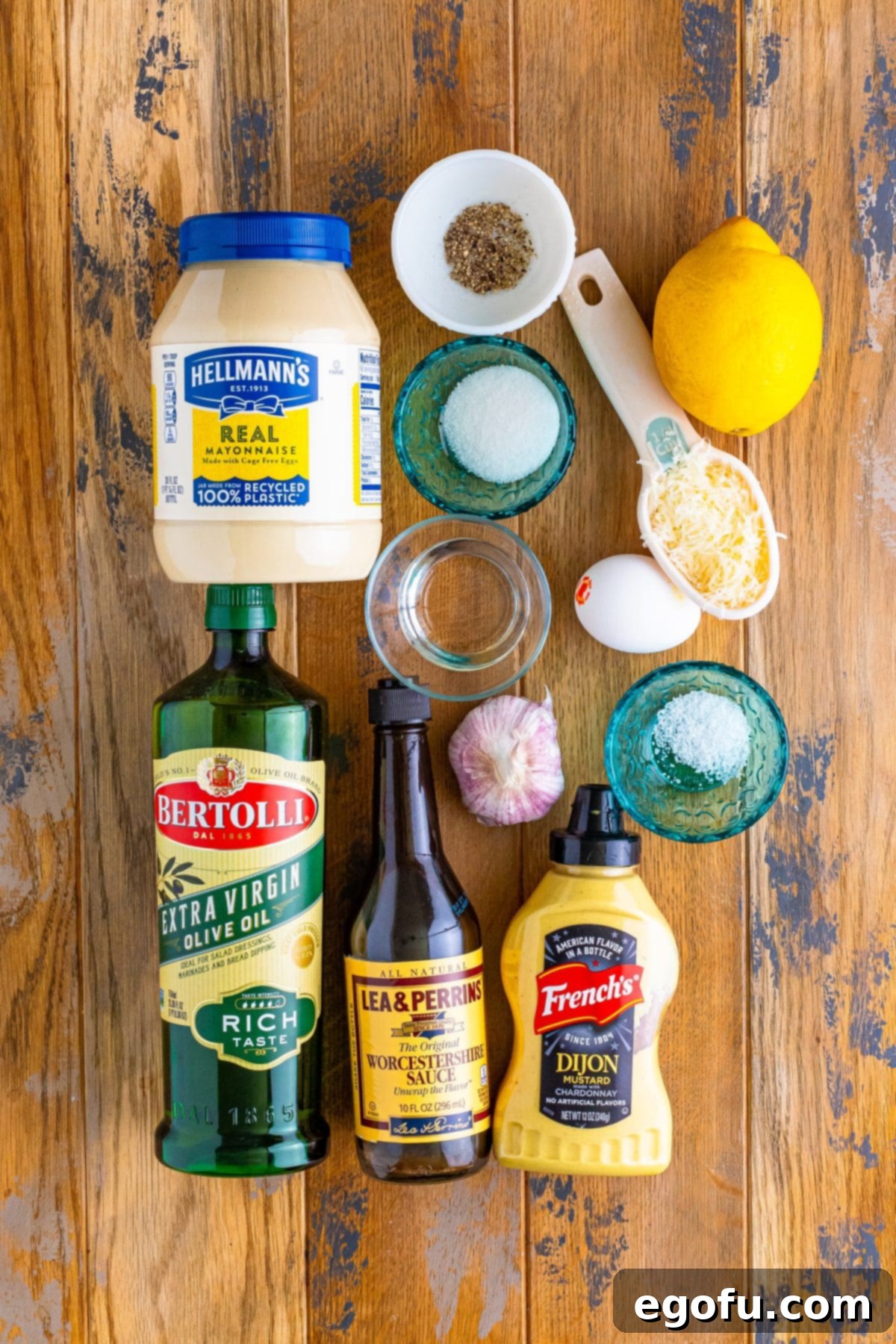 An array of fresh ingredients for Caesar salad dressing, including mayonnaise, lemon, olive oil, Worcestershire sauce, Dijon mustard, salt, pepper, garlic cloves, egg yolk, Parmesan cheese, water, and sugar, artfully arranged on a rustic surface.
