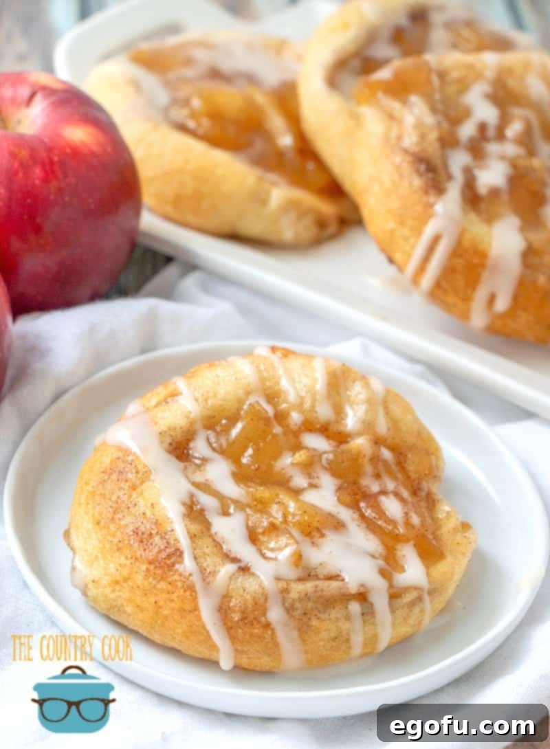 Golden Apple Danish Pastries 2 Crescent Roll Apple Danish on a white plate with a red apple in the background, highlighting its golden crust and delicious filling.