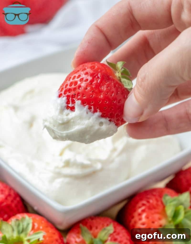A hand holding a whole strawberry dipped into a swirl of white creamy fruit dip, surrounded by a bounty of fresh, red strawberries, ready to be enjoyed.