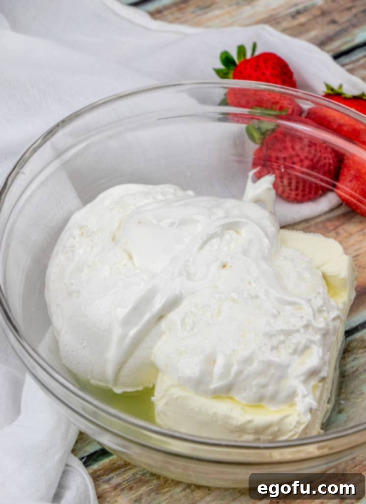 A clear bowl filled with the creamy fruit dip ingredients (cream cheese, marshmallow fluff, lemon juice) being mixed, with a side of fresh strawberries.