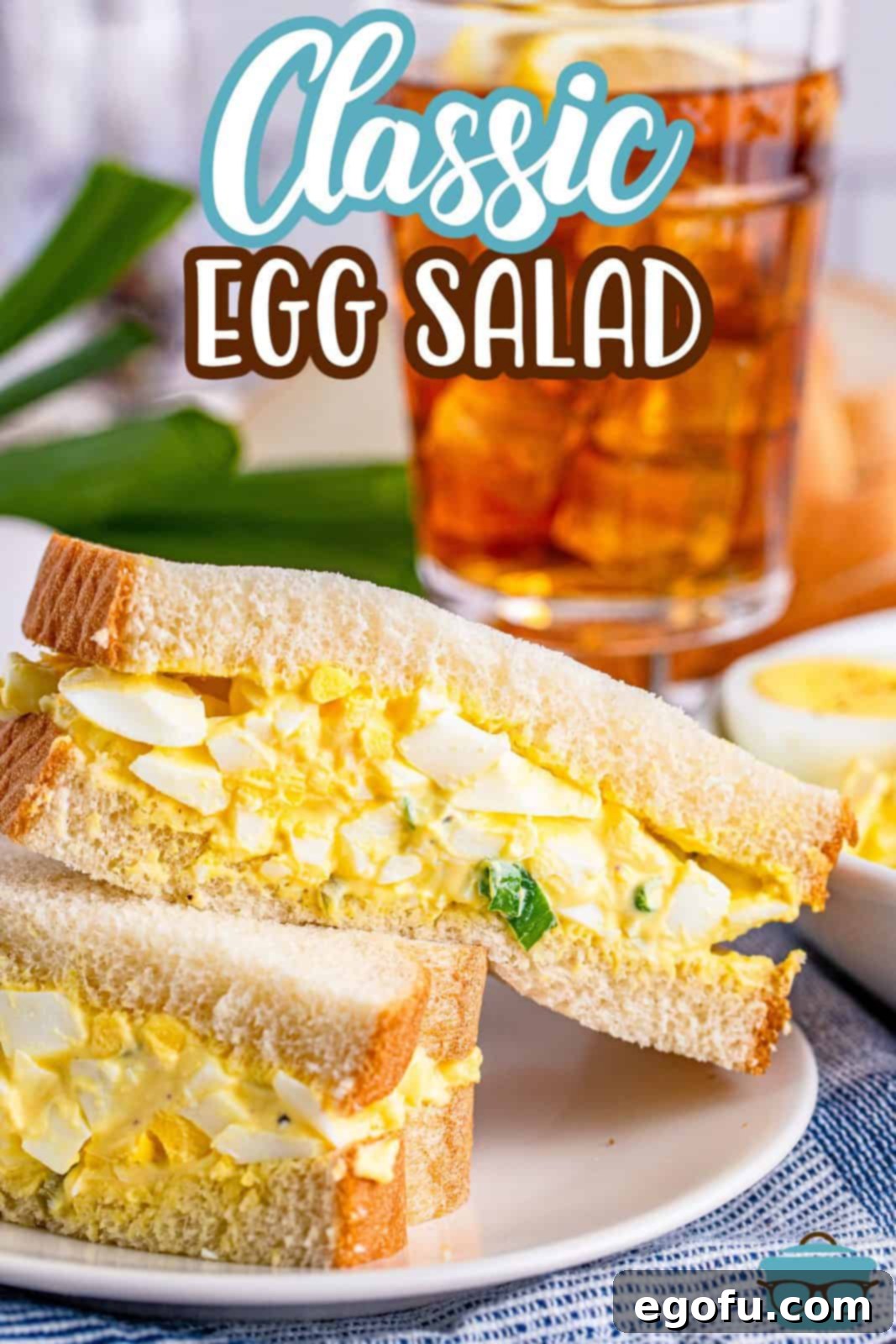 Irresistible Homemade Egg Salad 4 Two halves of an egg salad sandwich, showcasing the creamy filling, stacked on a white plate with a refreshing glass of iced tea in the background.