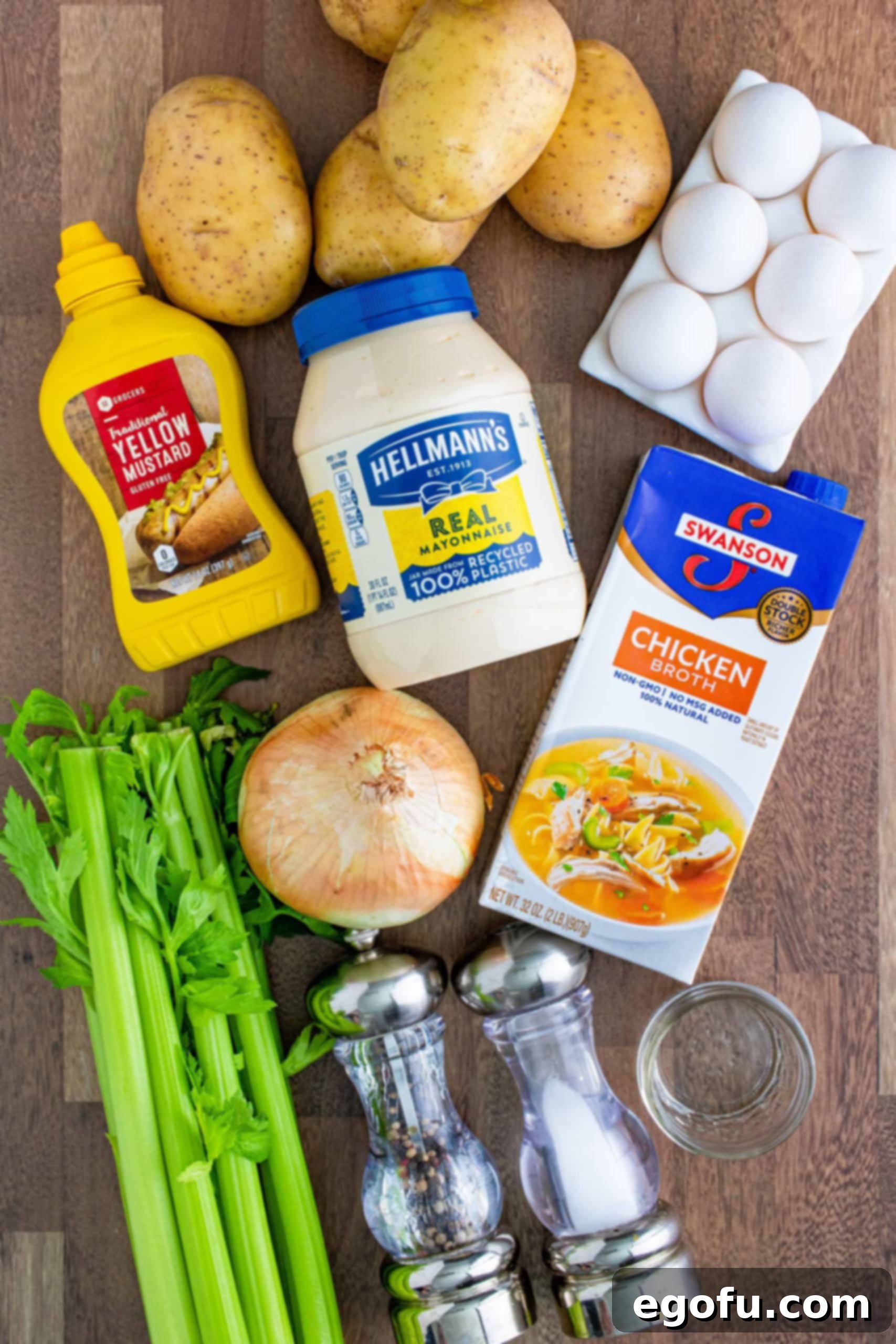 A beautifully arranged collage of fresh potato salad ingredients: Yukon gold potatoes, a jar of mayonnaise, white vinegar, yellow mustard, chicken stock, salt, pepper, celery stalks, a whole onion, and hard-boiled eggs.