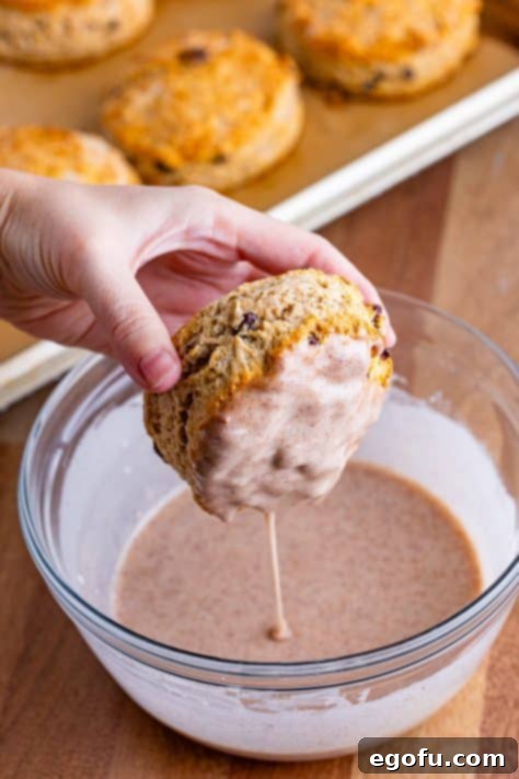 Buttery Cinnamon Raisin Biscuits 17 A hand dipping a biscuit in a cinnamon glaze.