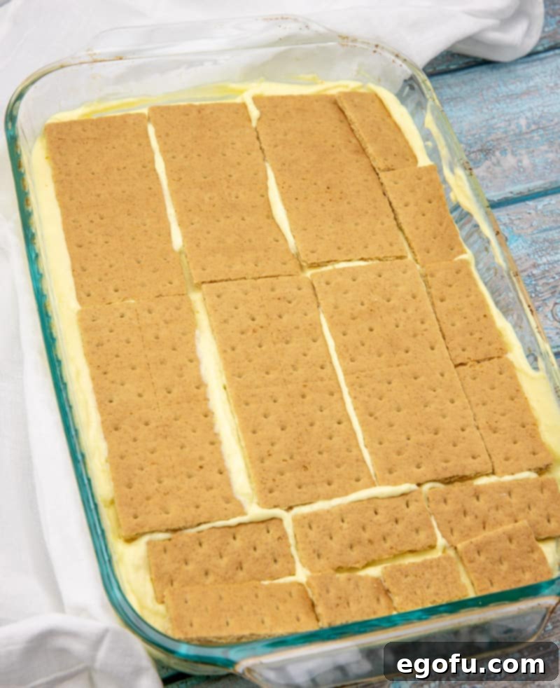 A baking dish showing distinct layers of graham crackers, vanilla pudding, and more graham crackers, ready for the next step.